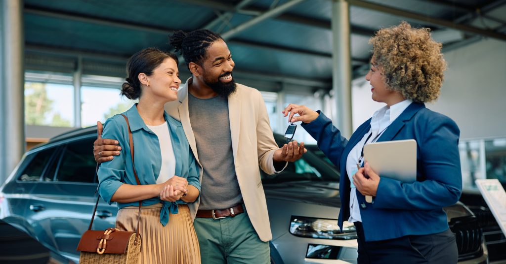 A salesperson handing over car keys to a couple with their car in the background